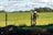A person wearing a hard hat and protective clothing is standing near a fence in a grassy field. The person appears to be talking on a phone. The background shows a wide expanse of green grass with some trees in the distance and a partly cloudy blue sky.