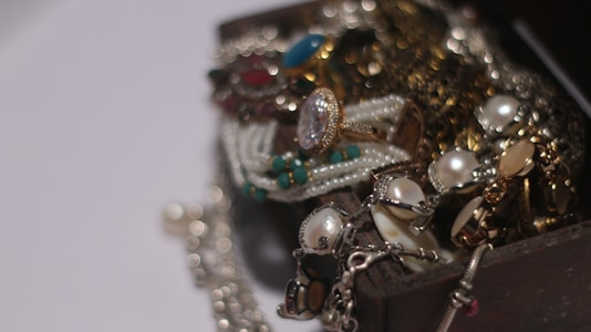 A collection of assorted jewelry spilling out of a wooden box, including pearl necklaces, gemstone rings, and gold chains. The arrangement creates a sense of luxury and abundance.