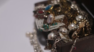 A collection of assorted jewelry spilling out of a wooden box, including pearl necklaces, gemstone rings, and gold chains. The arrangement creates a sense of luxury and abundance.