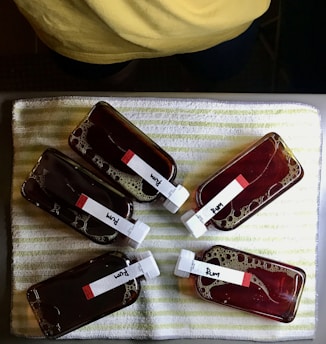 Five small, rectangular bottles filled with a dark liquid labeled 'Rum' are placed on a striped white and green towel. The bottles have white caps and are arranged in a circular pattern. Bubbles are visible within the liquid. Above the bottles, part of a person wearing a yellow garment is partially visible.