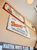 Several vintage-style advertisement signs are displayed on a wall. The signs feature brands such as Rudy's Farm Country Sausage, Elm Hill Meats, Shoney's Big Boy Restaurants, and Martha White Flour. Each sign has a retro design with unique fonts and illustrations, giving a nostalgic vibe.