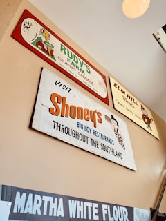 Several vintage-style advertisement signs are displayed on a wall. The signs feature brands such as Rudy's Farm Country Sausage, Elm Hill Meats, Shoney's Big Boy Restaurants, and Martha White Flour. Each sign has a retro design with unique fonts and illustrations, giving a nostalgic vibe.