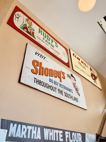 Several vintage-style advertisement signs are displayed on a wall. The signs feature brands such as Rudy's Farm Country Sausage, Elm Hill Meats, Shoney's Big Boy Restaurants, and Martha White Flour. Each sign has a retro design with unique fonts and illustrations, giving a nostalgic vibe.