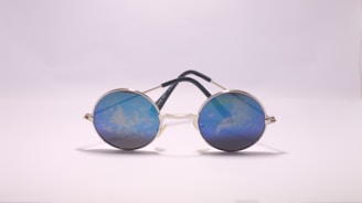 Round sunglasses with reflective blue lenses are placed on a flat surface, their metal frame supported by dark temples. The lenses reflect a cloudy pattern, giving a unique visual effect against the plain background.