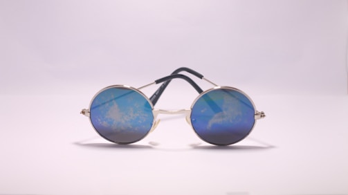 Round sunglasses with reflective blue lenses are placed on a flat surface, their metal frame supported by dark temples. The lenses reflect a cloudy pattern, giving a unique visual effect against the plain background.