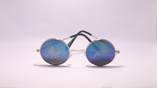 Round sunglasses with reflective blue lenses are placed on a flat surface, their metal frame supported by dark temples. The lenses reflect a cloudy pattern, giving a unique visual effect against the plain background.