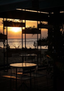 A serene beach scene during sunset, with the sun low on the horizon casting a golden glow over the ocean. Shadowy silhouettes of tables and chairs are visible in the foreground under an open-air structure adorned with plants, creating a peaceful atmosphere.