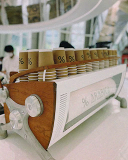 A row of brown paper coffee cups with a percentage symbol are neatly arranged on a wooden and metallic espresso machine. The background shows a modern, minimalistic interior with soft lighting and a blurred figure.