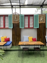 A cozy outdoor seating area features a wooden table with two metal chairs set on artificial grass. Behind, a cushioned bench is adorned with colorful pillows in red, yellow, and striped blue and white. Three birdcages hang above, in front of green shutters and wooden panels between red-framed windows under a translucent shelter.