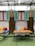 A cozy outdoor seating area features a wooden table with two metal chairs set on artificial grass. Behind, a cushioned bench is adorned with colorful pillows in red, yellow, and striped blue and white. Three birdcages hang above, in front of green shutters and wooden panels between red-framed windows under a translucent shelter.