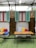 A cozy outdoor seating area features a wooden table with two metal chairs set on artificial grass. Behind, a cushioned bench is adorned with colorful pillows in red, yellow, and striped blue and white. Three birdcages hang above, in front of green shutters and wooden panels between red-framed windows under a translucent shelter.