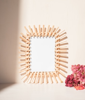 A decorative wooden frame with intricate patterns stands against a light background. To the side, a cluster of pink flowers adds a pop of color.