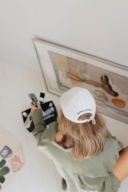 A person wearing a white cap with text on it leans over to observe a framed artwork placed on the floor. The individual has medium-length blonde hair and wears a green jacket. Nearby, they hold some photographs or documents in their hand.