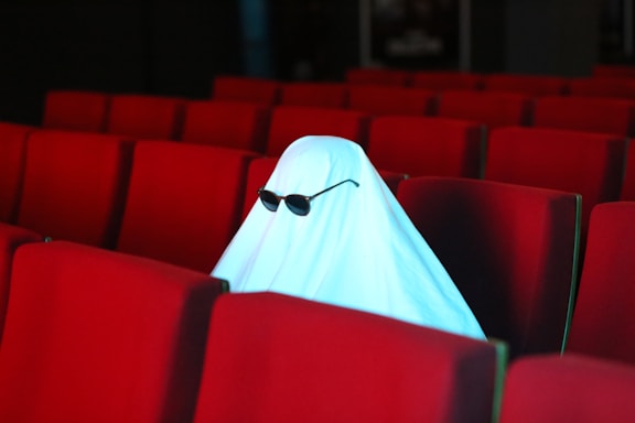 A figure dressed as a ghost sits in a theater filled with red, empty seats. The ghost costume is a simple white sheet with dark sunglasses worn over it.
