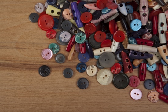 A collection of assorted buttons scattered on a wooden surface. The buttons come in various shapes, sizes, and colors, including circular and elongated styles. The vibrant colors include red, blue, yellow, green, pink, and beige tones, creating a visually striking composition.