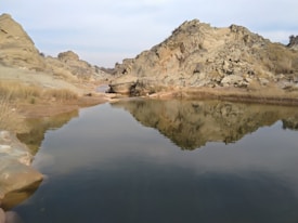 The image captures a serene natural landscape featuring a calm body of water reflecting the surrounding rocky hills. The terrain appears rugged with scattered dry grass, and the sky is overcast, contributing to a tranquil and quiet atmosphere.