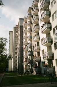 The image depicts a row of modern apartment buildings with multiple balconies. The buildings are white with dark railings, and some balconies have plants and flowers. The scene is set during daytime with partially cloudy skies. In the foreground, there are several bicycles parked on a paved path alongside a grassy area with trees.