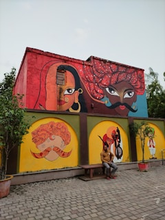 A building wall adorned with colorful, traditional Indian mural art. The murals feature stylized human faces with elaborate turbans and facial features. A man is seated on a bench in front of the wall, which is bordered by small trees in pots.