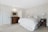 A cozy bedroom features a large bed with white and floral bedding, flanked by two wooden nightstands topped with matching lamps. A wooden dresser with ornate details sits against a light-colored paneled wall, and a painting hangs above it. The room is softly lit, with a white door on one side.
