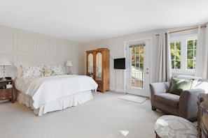 A spacious and well-lit bedroom features a large bed with patterned pillows and a neutral bedspread. There are two bedside tables with lamps on each side of the bed. An elegant wooden wardrobe is placed against one wall, adjacent to a flat-screen television mounted nearby. The room includes a comfortable armchair with green cushions next to a window draped with light-colored curtains. A door leads to a balcony, inviting natural light to flood the room.