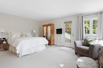 A spacious and well-lit bedroom features a large bed with patterned pillows and a neutral bedspread. There are two bedside tables with lamps on each side of the bed. An elegant wooden wardrobe is placed against one wall, adjacent to a flat-screen television mounted nearby. The room includes a comfortable armchair with green cushions next to a window draped with light-colored curtains. A door leads to a balcony, inviting natural light to flood the room.