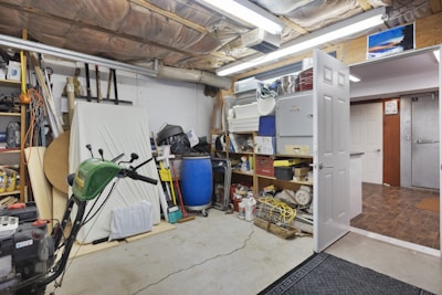 A cluttered garage space with various tools and equipment scattered around. A green lawnmower is visible in the foreground, surrounded by shelves filled with boxes, bottles, and other items. A blue barrel and cleaning supplies are in the corner, while a partially open door leads to an adjacent room.