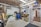 A cluttered garage space with various tools and equipment scattered around. A green lawnmower is visible in the foreground, surrounded by shelves filled with boxes, bottles, and other items. A blue barrel and cleaning supplies are in the corner, while a partially open door leads to an adjacent room.
