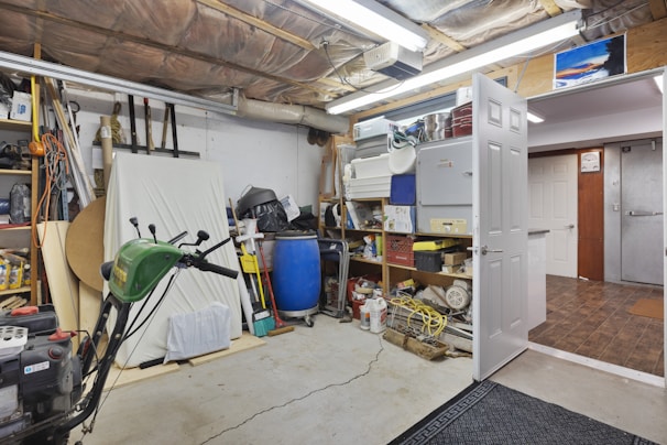 A cluttered garage space with various tools and equipment scattered around. A green lawnmower is visible in the foreground, surrounded by shelves filled with boxes, bottles, and other items. A blue barrel and cleaning supplies are in the corner, while a partially open door leads to an adjacent room.