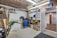 A cluttered garage space with various tools and equipment scattered around. A green lawnmower is visible in the foreground, surrounded by shelves filled with boxes, bottles, and other items. A blue barrel and cleaning supplies are in the corner, while a partially open door leads to an adjacent room.