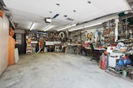 A cluttered workshop with various tools and equipment neatly organized on shelves and workbenches. The room is well-lit with overhead fluorescent lights, and the floor is concrete. There are items like ladders, power tools, and mechanical parts visible throughout the space.