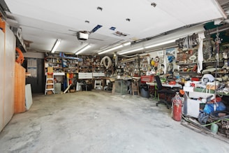 A cluttered workshop with various tools and equipment neatly organized on shelves and workbenches. The room is well-lit with overhead fluorescent lights, and the floor is concrete. There are items like ladders, power tools, and mechanical parts visible throughout the space.