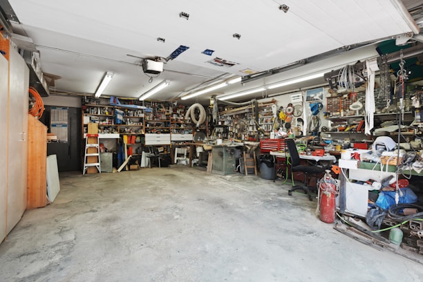 A cluttered workshop with various tools and equipment neatly organized on shelves and workbenches. The room is well-lit with overhead fluorescent lights, and the floor is concrete. There are items like ladders, power tools, and mechanical parts visible throughout the space.