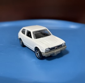 A small white toy car is placed on a smooth blue surface. The car features detailed front headlights, a distinct grille, and black tires with silver rims. Its compact hatchback design is reminiscent of vintage models.