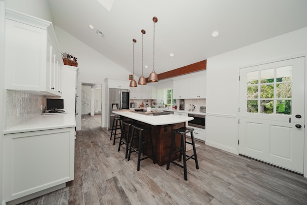A modern kitchen features white cabinetry with a central island, equipped with black stools. Pendant lights in a row hang above the island, and stainless steel appliances are integrated into the cabinetry. The floor is made of wood-like material, and there is a door with glass panels leading outside. A window allows natural light to illuminate the space.