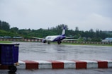 A stationary airplane with a purple and white livery is positioned on a wet airport runway with lush green hills and trees in the background. The ground is wet from rain, reflecting the aircraft slightly. In the foreground, a blue baggage cart is visible, bordered by red and white striped barriers.
