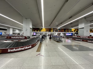A spacious airport baggage claim area with multiple conveyor belts, adorned with advertising banners. Several people are seen collecting luggage, and there are carts available for baggage. The area is well-lit with overhead lighting, and the floor is tiled with a clean, polished look.
