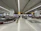 A spacious airport baggage claim area with multiple conveyor belts, adorned with advertising banners. Several people are seen collecting luggage, and there are carts available for baggage. The area is well-lit with overhead lighting, and the floor is tiled with a clean, polished look.