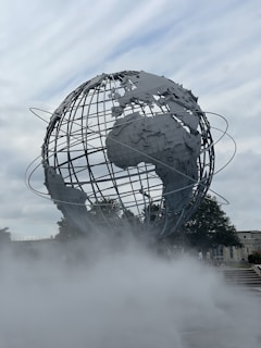 A large, metallic globe structure with visible continents is surrounded by mist, set against a cloudy sky. The globe is encircled by thin metal rings representing orbital paths. Trees and a building can be seen in the background.