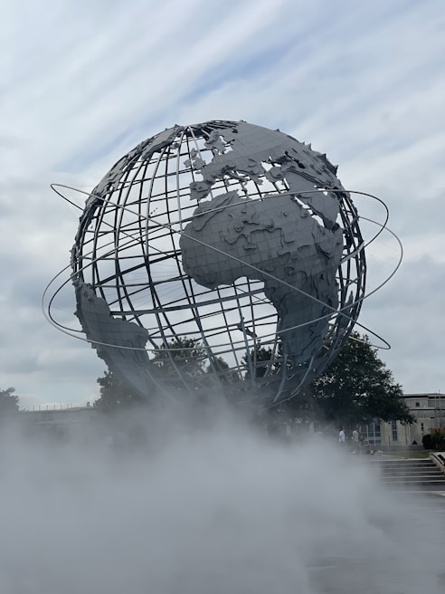 A large, metallic globe structure with visible continents is surrounded by mist, set against a cloudy sky. The globe is encircled by thin metal rings representing orbital paths. Trees and a building can be seen in the background.