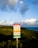 A warning sign stands prominently in the foreground, set against a backdrop of lush greenery and an expansive ocean view. The sky above is filled with fluffy, scattered clouds, illuminated by soft, ambient light. The sign contains warnings about the area, possibly indicating natural hazards.