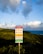 A warning sign stands prominently in the foreground, set against a backdrop of lush greenery and an expansive ocean view. The sky above is filled with fluffy, scattered clouds, illuminated by soft, ambient light. The sign contains warnings about the area, possibly indicating natural hazards.