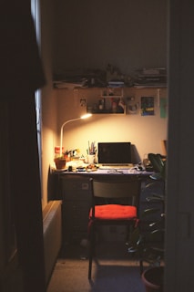 A cozy, dimly-lit workspace with a wooden desk cluttered with papers, pens, and a laptop. A warm lamp casts a soft glow over the area, highlighting a potted plant and a chair with a red cushion. The walls are covered with shelves holding various items and artwork.