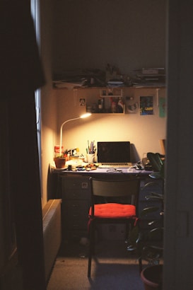 A cozy, dimly-lit workspace with a wooden desk cluttered with papers, pens, and a laptop. A warm lamp casts a soft glow over the area, highlighting a potted plant and a chair with a red cushion. The walls are covered with shelves holding various items and artwork.