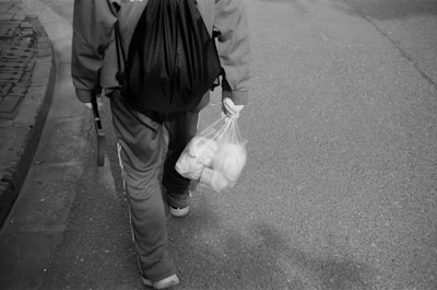 A person wearing a tracksuit and carrying a backpack walks along a street. In their left hand, they hold a plastic bag filled with groceries or items. The scene is in black and white, capturing a moment of daily life.