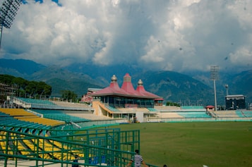 A large cricket stadium with an empty field is set against a backdrop of majestic mountains with swirling clouds. The stadium's architecture features a striking red roof with round decorative elements. Empty seats with a mix of yellow and green can be seen in the stands. Tall floodlights tower over the field, and lush green trees surround parts of the stadium.