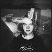 A black and white portrait of a person with long hair, wearing a dark top with a graphic design. In the background, there is a blurred scene of a room with indistinct objects like cups and utensils, giving a layered, double exposure effect.
