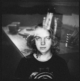 A black and white portrait of a person with long hair, wearing a dark top with a graphic design. In the background, there is a blurred scene of a room with indistinct objects like cups and utensils, giving a layered, double exposure effect.