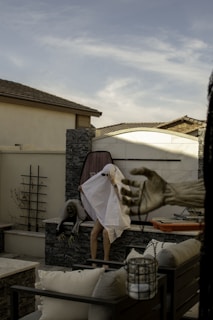 A person draped with a white sheet and sunglasses is standing on a patio with Halloween decorations, including a ghoul figure and a skeletal hand reaching from the right. Outdoor furniture is visible in the foreground and background under a partly cloudy sky.