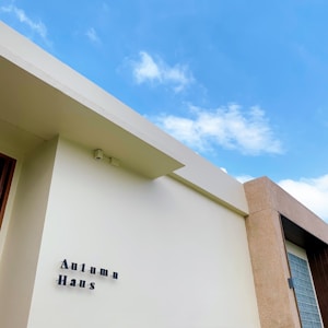 A modern building facade with a minimalist design featuring clean lines. The text 'Autumn Haus' is displayed on the light-colored wall. The sky is bright and blue with a few scattered clouds.