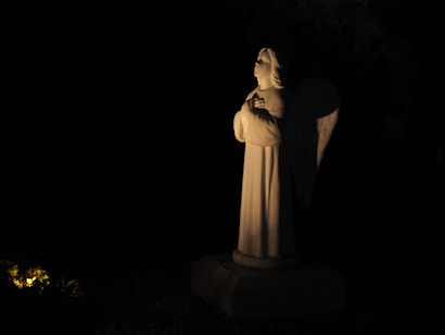 A white marble statue of an angel stands illuminated against a dark background. The angel has wings and is depicted with its hands clasped in a prayerful pose. Soft lighting casts gentle shadows, emphasizing the serene and contemplative expression on its face. There is a small area with yellow lighting and greenery at the bottom left corner.